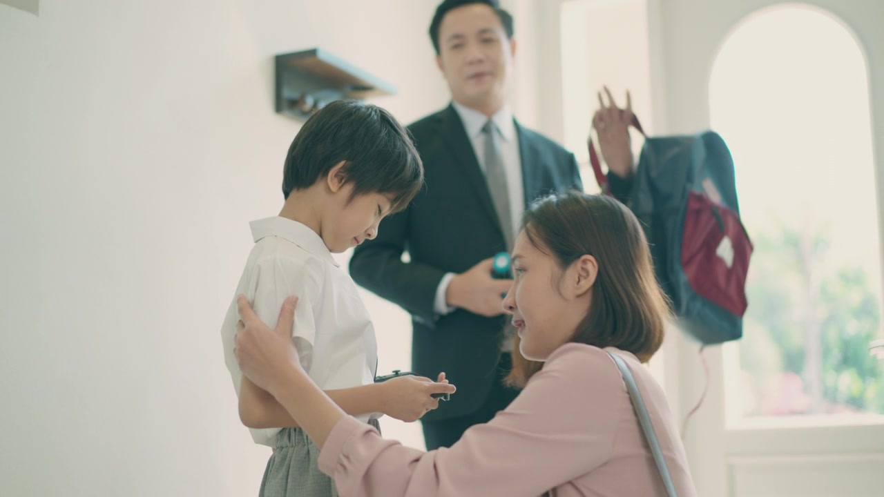 詳情圖-亞洲媽媽抱著她的小兒子，準(zhǔn)備送他去上學(xué)，而亞洲爸爸在家里的客廳里背著兒子的書(shū)包，準(zhǔn)備去上學(xué)。親子關(guān)系或愛(ài)與紐帶表達(dá)的概念。