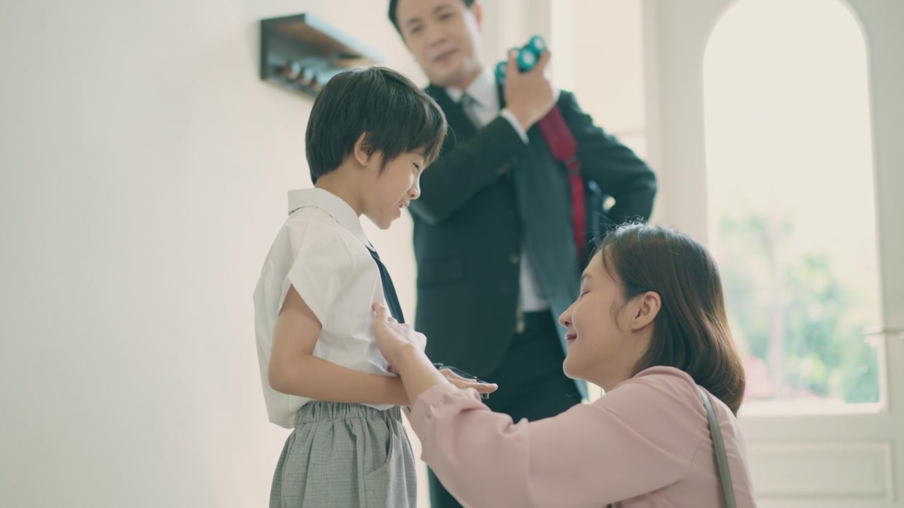 詳情圖-亞洲媽媽抱著她的小兒子，準(zhǔn)備送他去上學(xué)，而亞洲爸爸在家里的客廳里背著兒子的書(shū)包，準(zhǔn)備去上學(xué)。親子關(guān)系或愛(ài)與紐帶表達(dá)的概念。