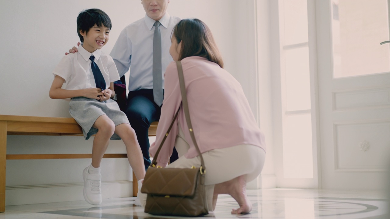 6岁的亚洲小男孩和他英俊的父亲坐在长椅上，父亲正在为儿子准备上学的书包，而亚洲母亲正在帮儿子系鞋带。早上学校的例行公事。