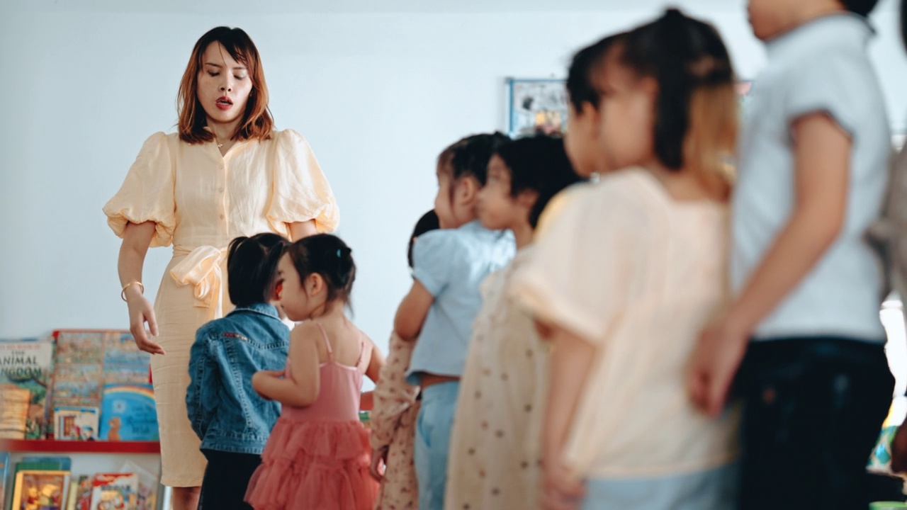 亚裔蒙台梭利幼教老师早上引导并安排排队进入教室的学生
