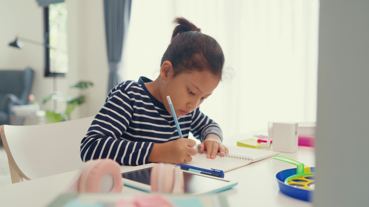 亚洲蹒跚学步的女孩与毛衣坐在书桌前与笔记本使用铅笔专注写笔记本做作业从网上学习课程在周末在家。