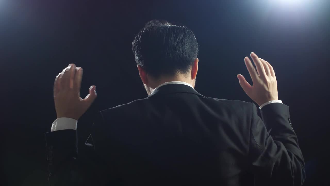 在黑屏演播室讲话时，穿着西装的亚洲演讲者竖起食指的特写背影