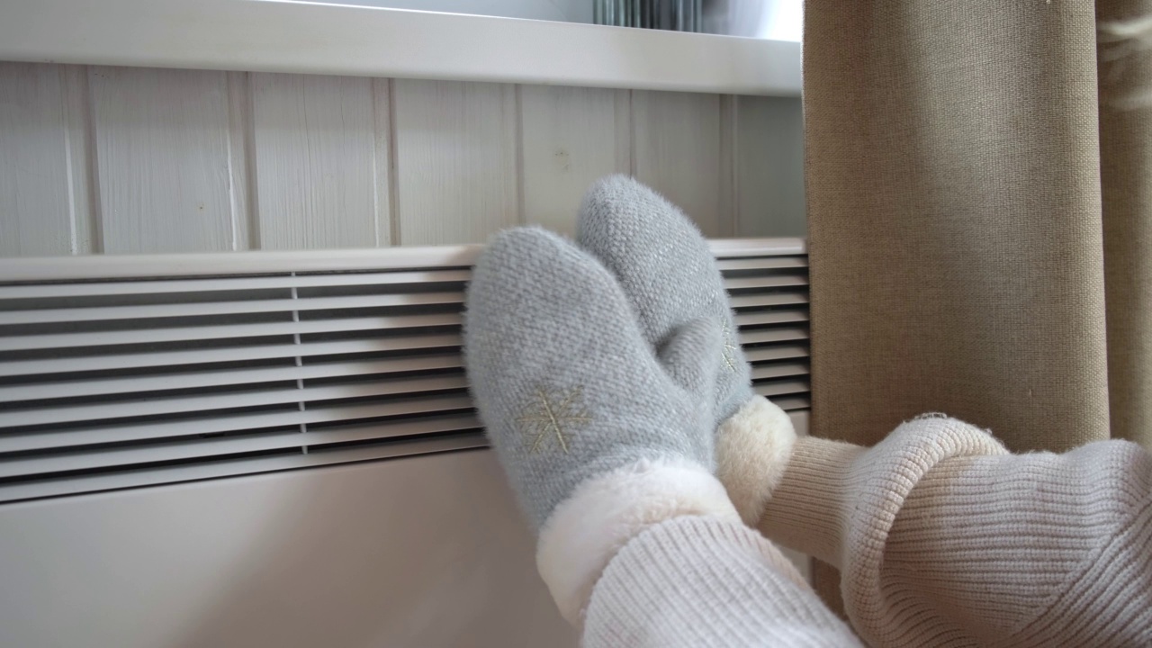 手戴着冬天的手套，在电加热器上加热。被冻住的女人戴着针织连指手套冻得严严实实。不舒服的呆在家里。女孩在现代暖气片上暖手。