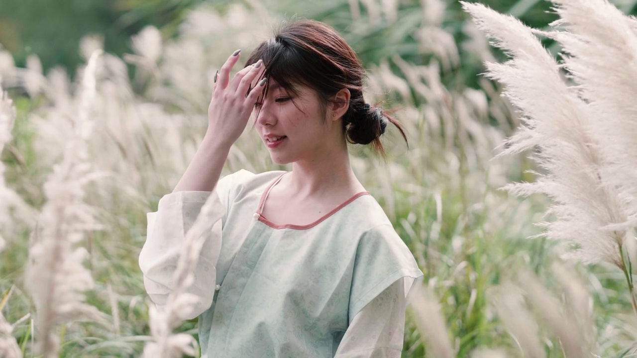 美麗的中國女孩穿著傳統(tǒng)的衣服，在秋日的蘆葦叢中享受微風(fēng)，閉上眼睛微笑著看著相機，4k慢鏡頭。