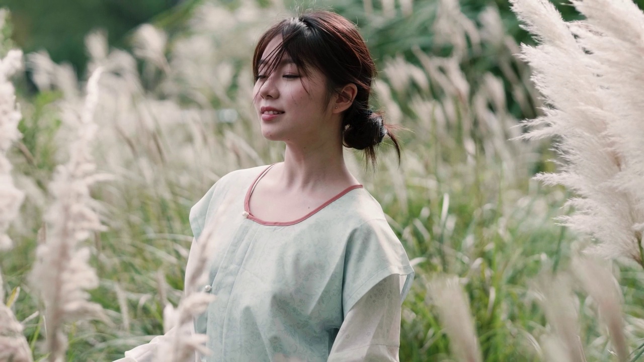 詳情圖-美麗的中國女孩穿著傳統(tǒng)的衣服，在秋日的蘆葦叢中享受微風(fēng)，閉上眼睛微笑著看著相機(jī)，4k慢鏡頭。