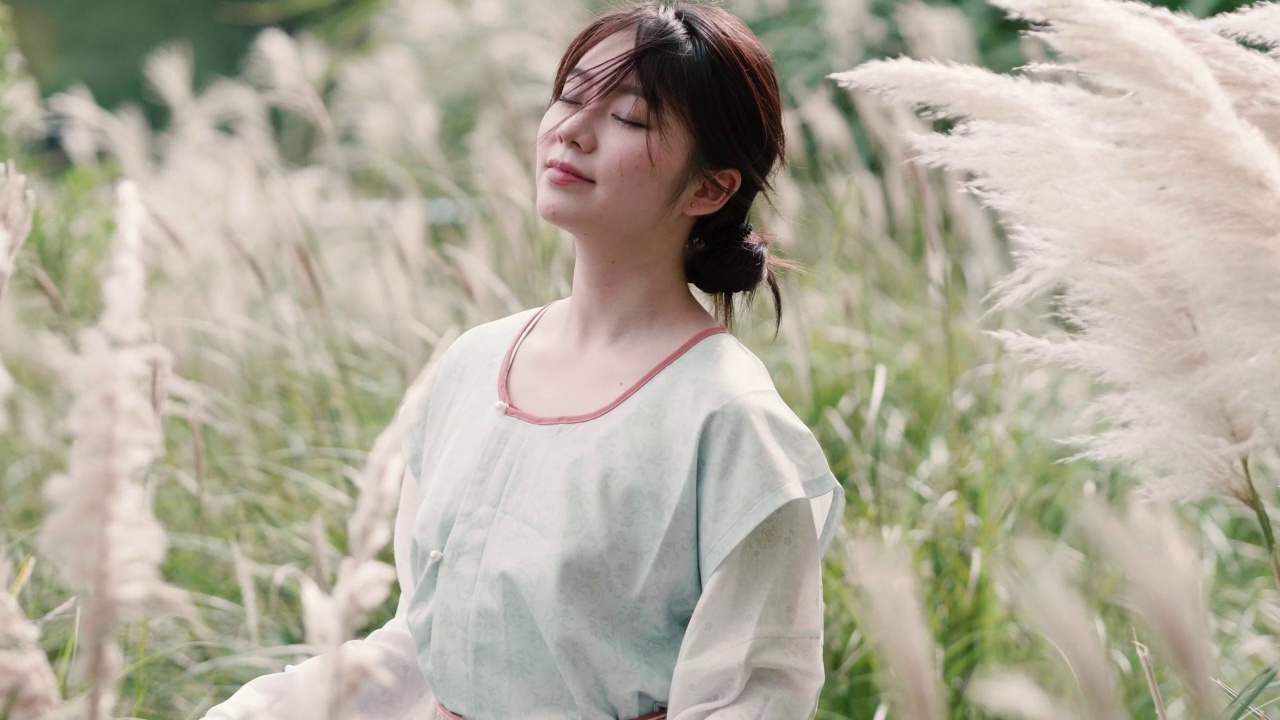 詳情圖-美麗的中國女孩穿著傳統(tǒng)的衣服，在秋日的蘆葦叢中享受微風(fēng)，閉上眼睛微笑著看著相機(jī)，4k慢鏡頭。