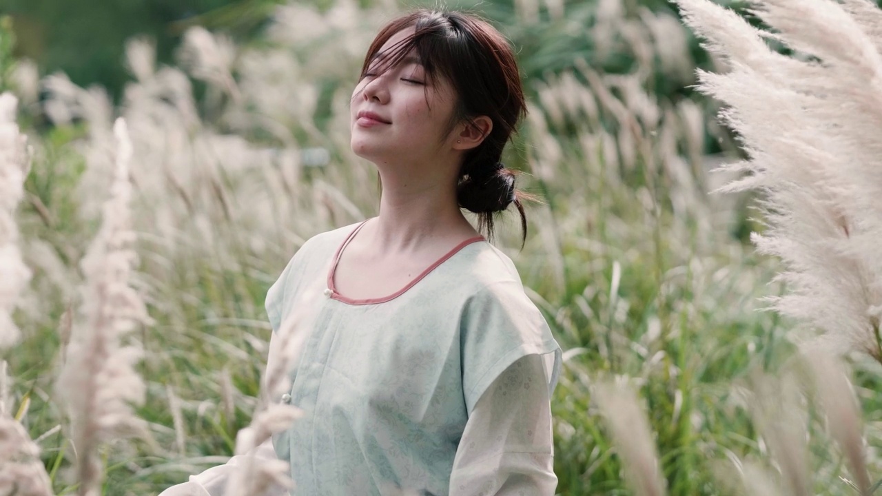 詳情圖-美麗的中國女孩穿著傳統(tǒng)的衣服，在秋日的蘆葦叢中享受微風(fēng)，閉上眼睛微笑著看著相機(jī)，4k慢鏡頭。