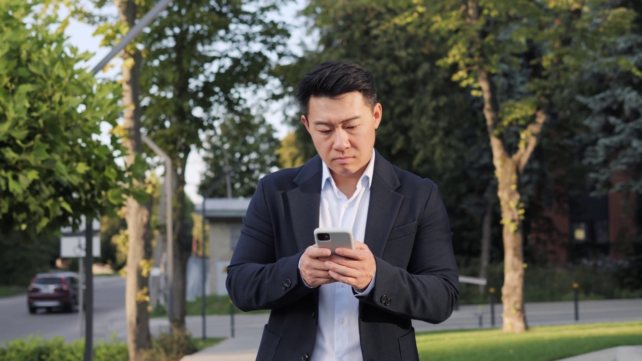 亚洲专注的商人走在城市街道城市附近的办公中心使用手机短信滚动点击屏幕手机，新技术通信晴天成功慢动作。