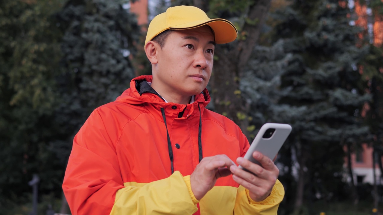 专注的亚洲快递员在他的智能手机上写短信，使用系统导航寻找建筑。有人在户外用智能手机敲击或发短信。男快递员正在打电话。