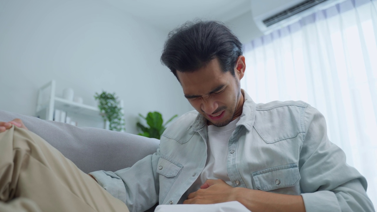 亚洲有魅力的病男在客厅痛得肚子疼。英俊的年轻人坐在沙发上，肚子疼，肚子疼，然后在房子里抱着肚子。健康问题