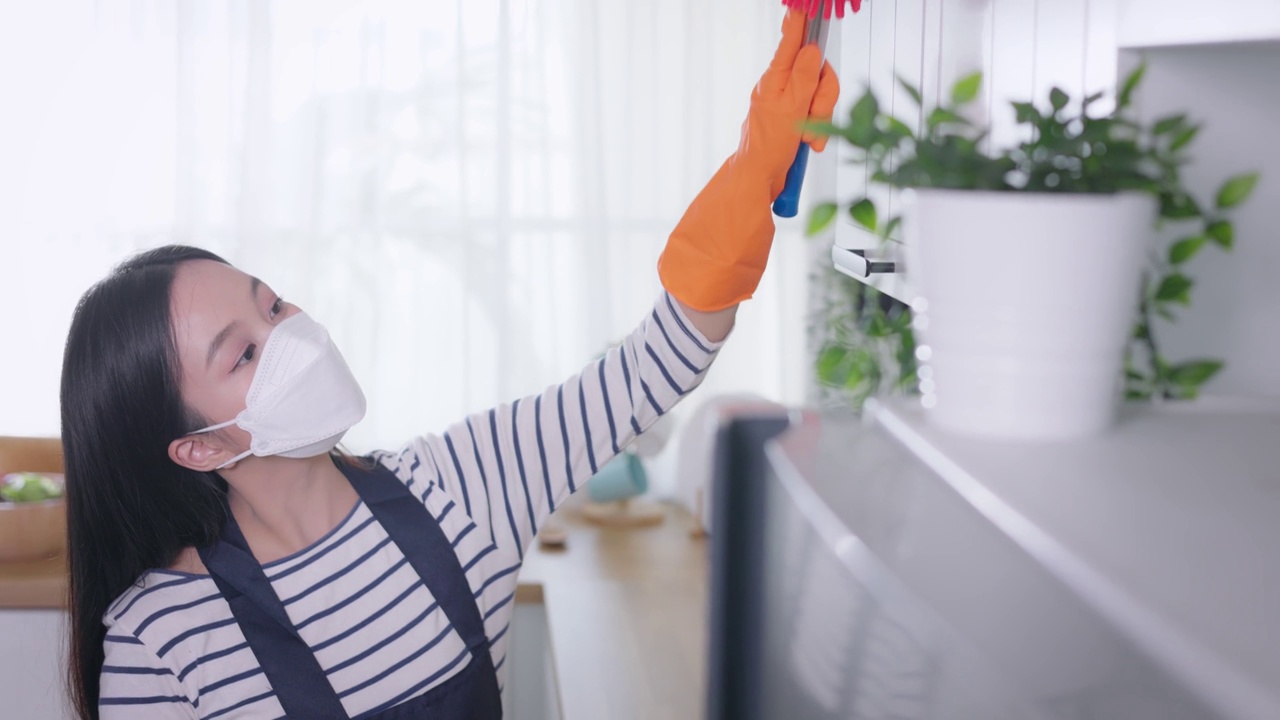年轻的亚洲妇女用掸子打扫家。美丽的清洁服务人员打扫卫生和整理厨房。家务和杂务概念