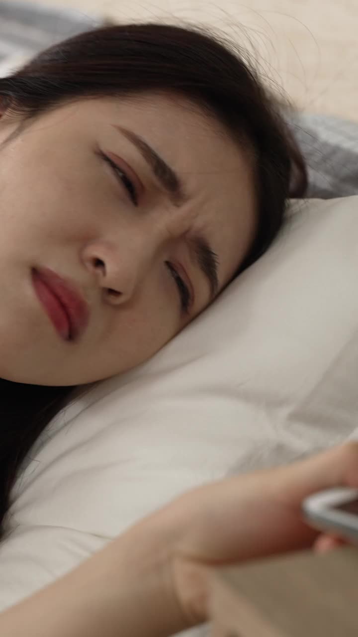 垂直屏幕:近距离观察一个昏昏欲睡的亚洲女性，她的手机被闹钟唤醒，关机后，在家里的卧室查看时间，她仍然在睡觉。