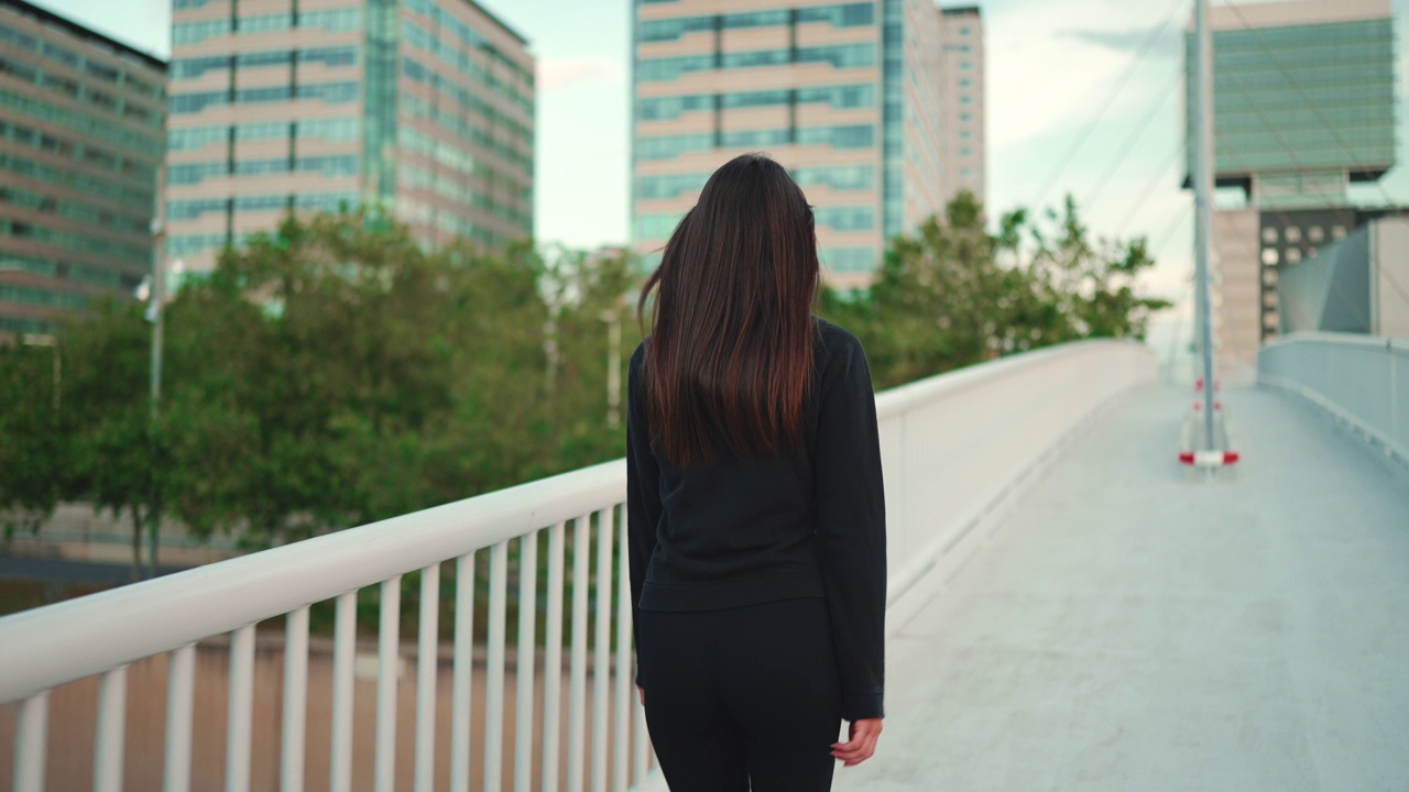 年轻女子披头散发，穿着黑色运动帽衫，正快步走过现代都市桥面。后视图