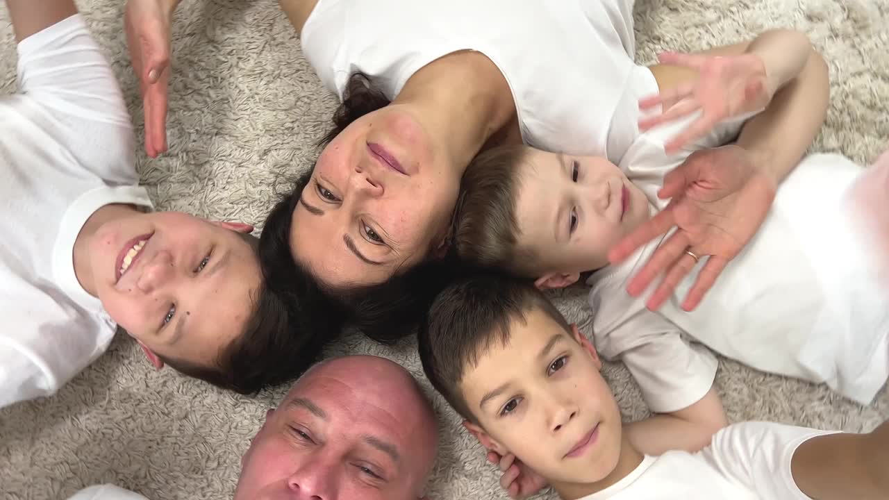 快乐快乐的一家人躺在地毯上，面朝上，微笑着三个男孩，兄弟，妈妈，爸爸，爸爸，男人，光头，女人，都穿着白色t恤，快乐的家庭度假，舒适的女人，最后咳嗽