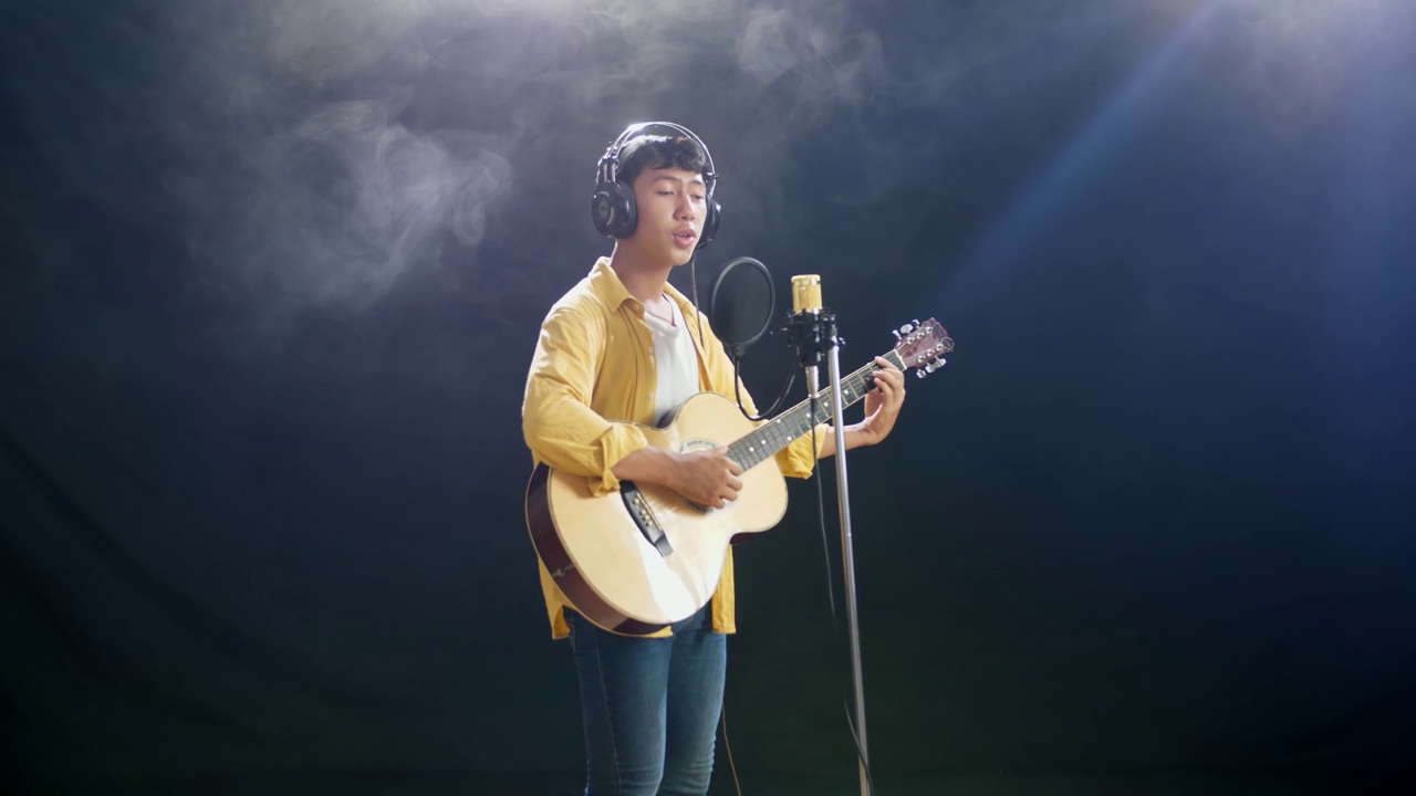侧视图年轻的亚洲男孩戴着耳机弹吉他，对着电容麦克风在白烟黑色背景上唱歌