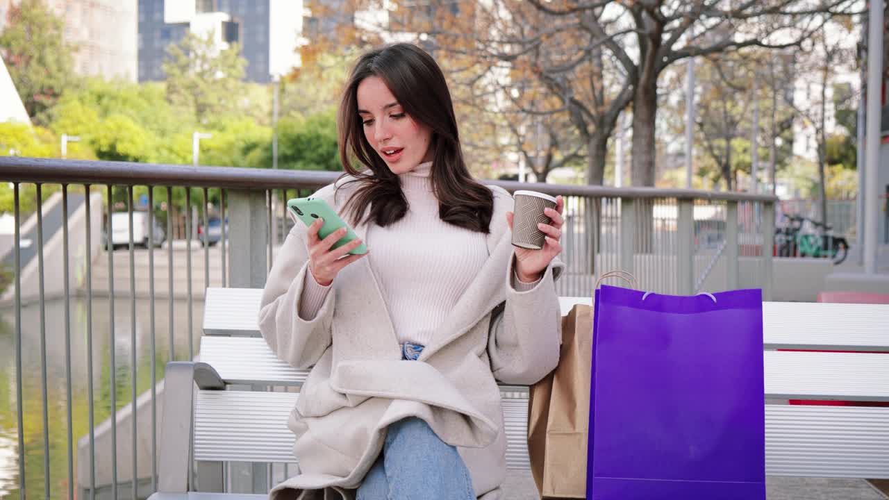 一位黑发女士购物后坐在城市街道的长凳上用手机打电话。孤僻的年轻女人。微笑着用智能手机聊天。通信的概念。慢动作