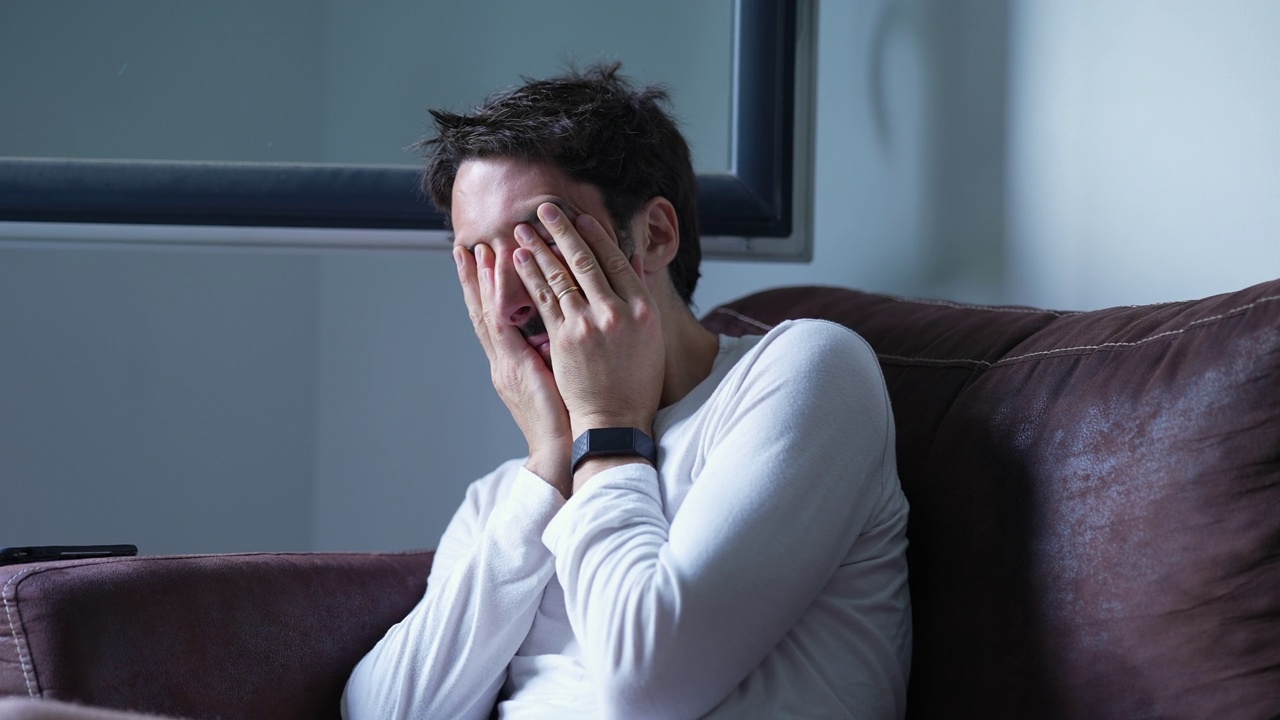 压力大的男人揉脸和眼睛，坐在家里的沙发上感到精神压力。沮丧的人用手揉眼睛