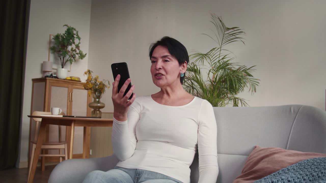 快乐美丽的55-60岁的老年妇女坐在家里的沙发上和家人聊天，用智能手机视频电话飞吻。