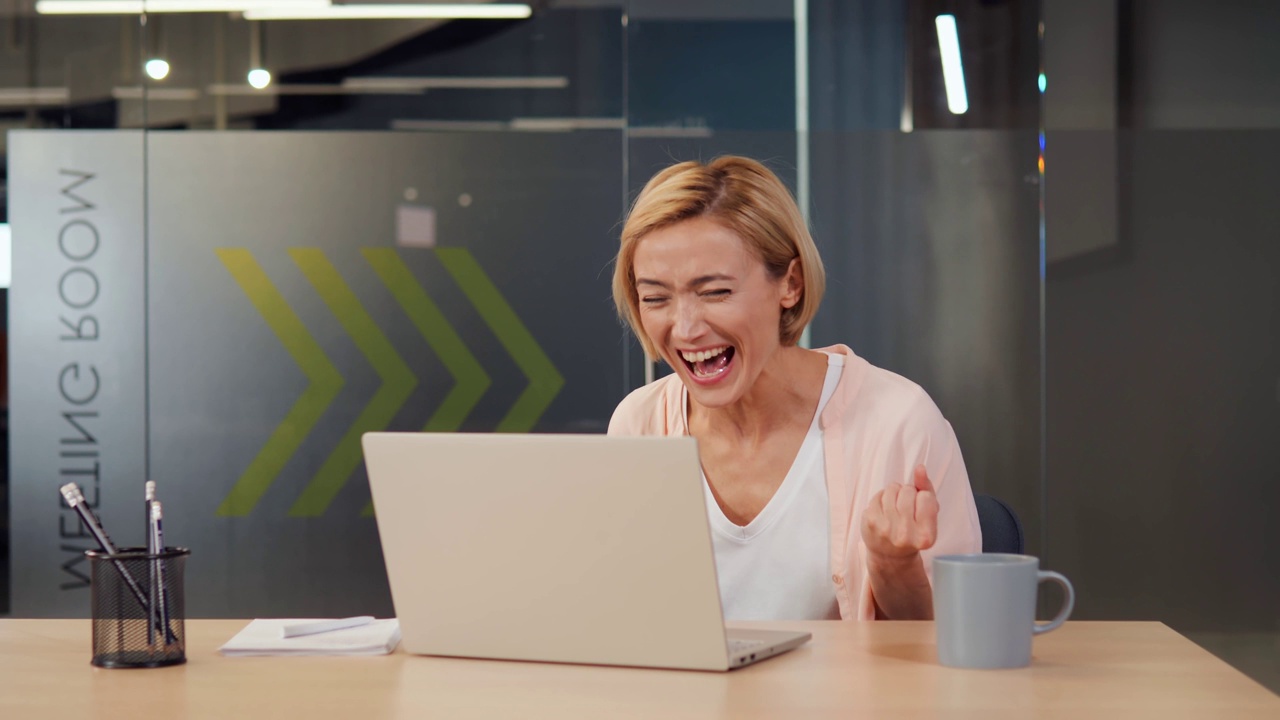 詳情圖-美麗，開朗，精力充沛的女人在她的筆記本電腦上收到一個獲勝的通知。女孩因成功和勝利而欣喜若狂。她燦爛的笑容和正能量可以照亮整個房間。
