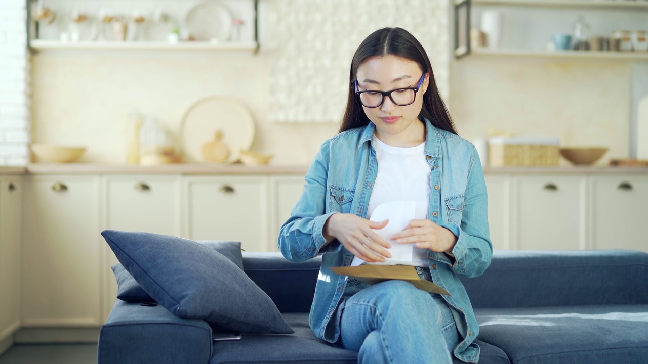 年轻微笑的亚洲女子坐在客厅的沙发上读着带着好消息的信。