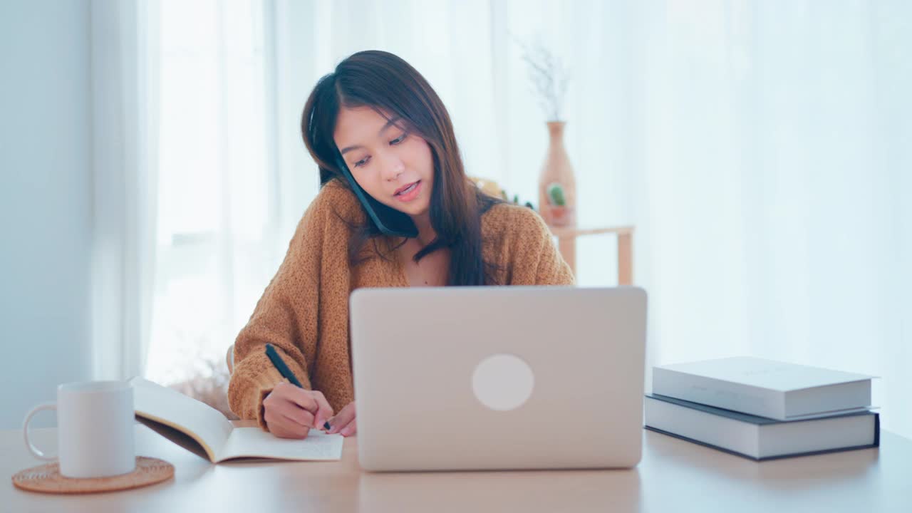 年轻忙碌的亚洲女性在笔记本电脑上做项目，在移动智能手机上与公司客户或同事交谈，在纸上做笔记，在家工作。