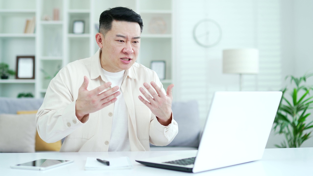 愤怒兴奋的亚洲商人坐在家里办公室用笔记本电脑争论视频通话。