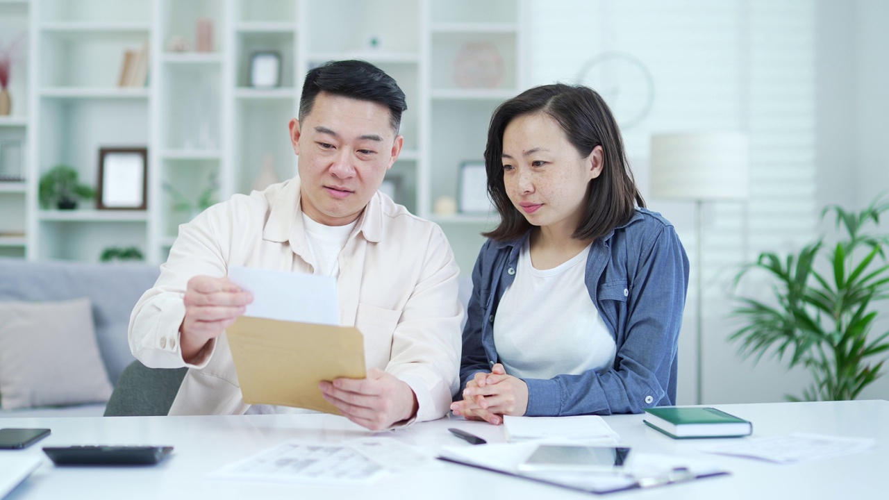 幸福的亚洲夫妇庆祝成功，坐在家里办公室读了一封带着好消息的信