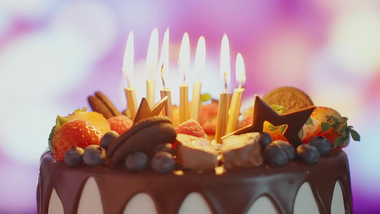 以散景灯泡花环为背景，为孩子生日派对准备的明亮甜点。生日蛋糕与燃烧的蜡烛，巧克力釉是旋转的站在桌子上的房子。家庭。童年