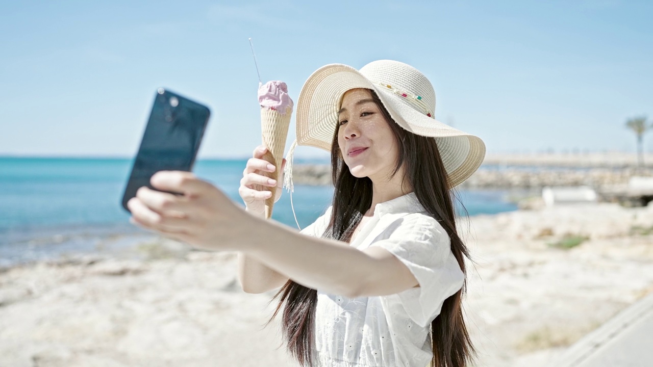 年轻的中国女游客在海边边吃冰淇淋边用智能手机自拍