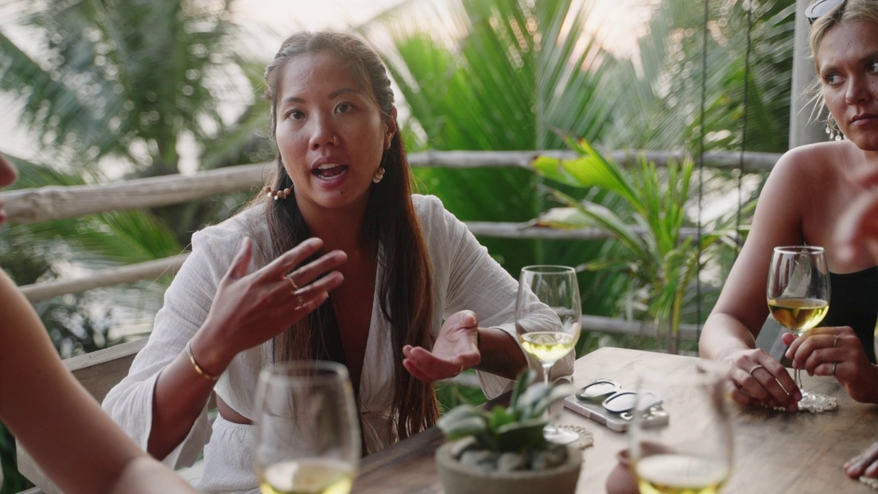 不同的女性朋友在日落的屋顶海景餐厅喝酒。黄昏时分，多种族妇女在热带咖啡馆里谈笑风生。年轻的多民族妇女在用餐。棕榈树，背景是海洋。