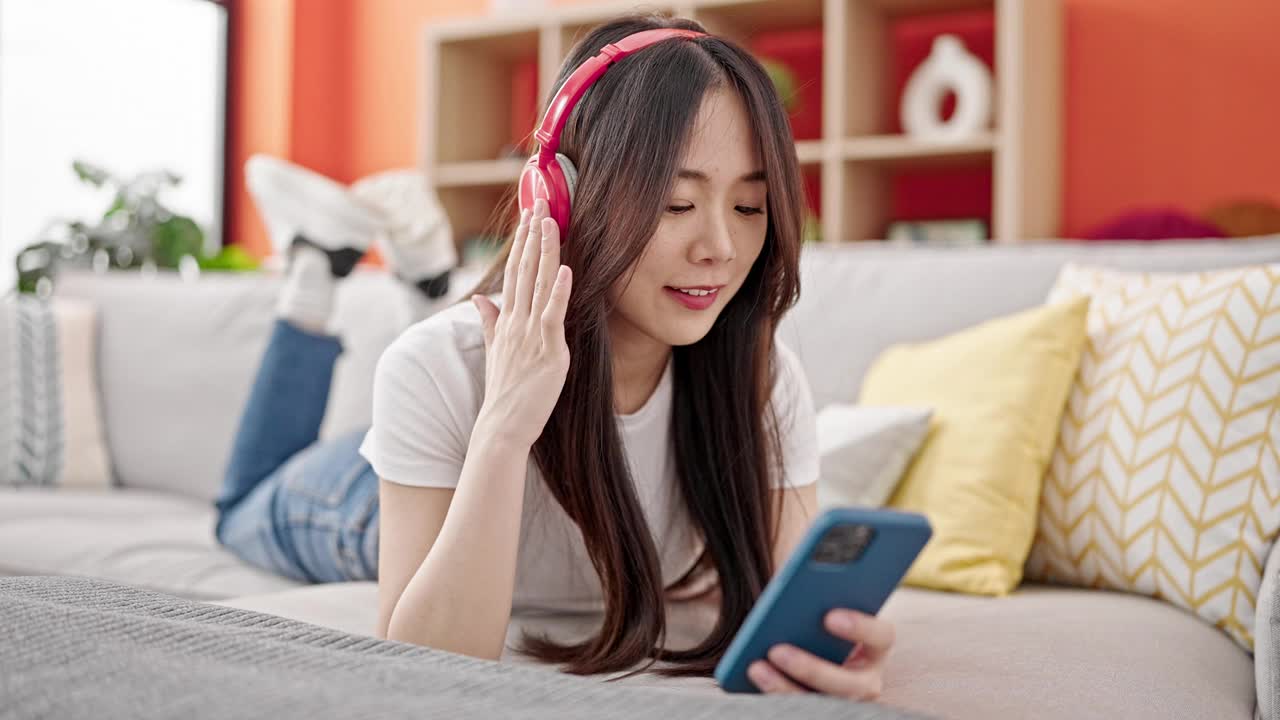 年轻的中国女子戴着耳机躺在家里的沙发上玩智能手机