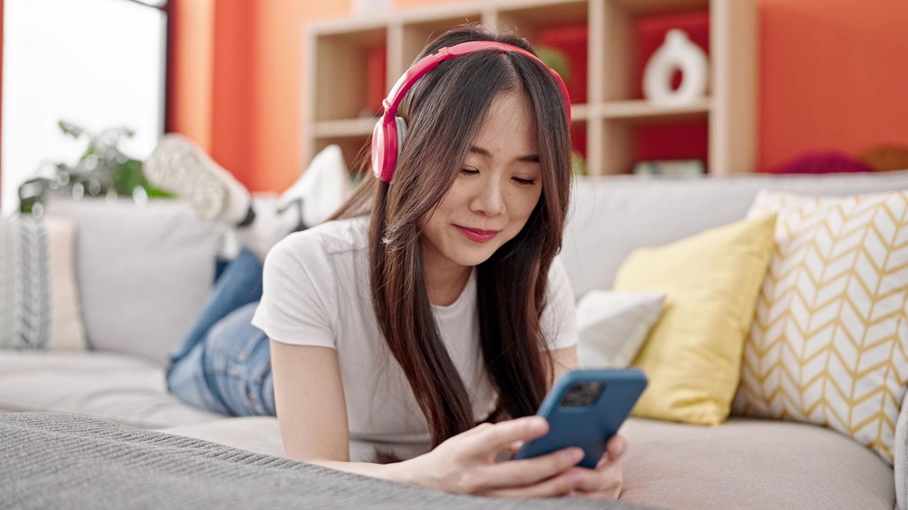 詳情圖-年輕的中國女子戴著耳機(jī)躺在家里的沙發(fā)上玩智能手機(jī)