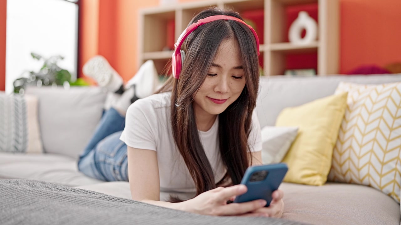 詳情圖-年輕的中國女子戴著耳機(jī)躺在家里的沙發(fā)上玩智能手機(jī)