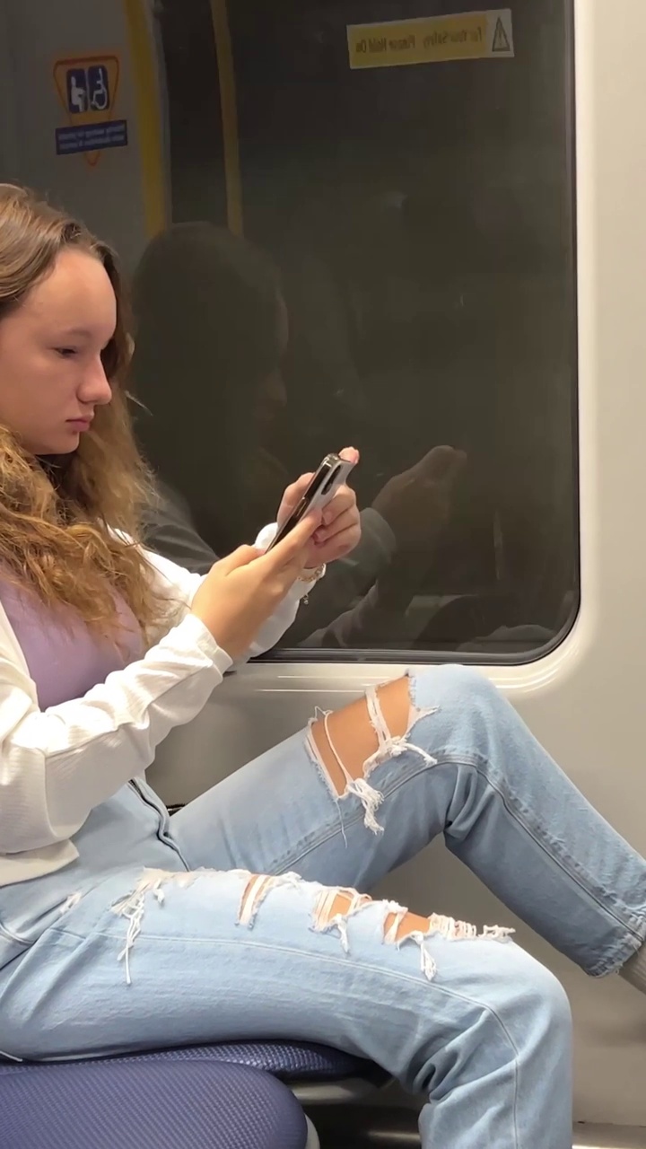 乌克兰金发的欧洲女孩坐在轻轨上，她在她面前听音乐，拿着手机，她穿着白色衬衫和蓝色破洞牛仔裤，她在隧道里