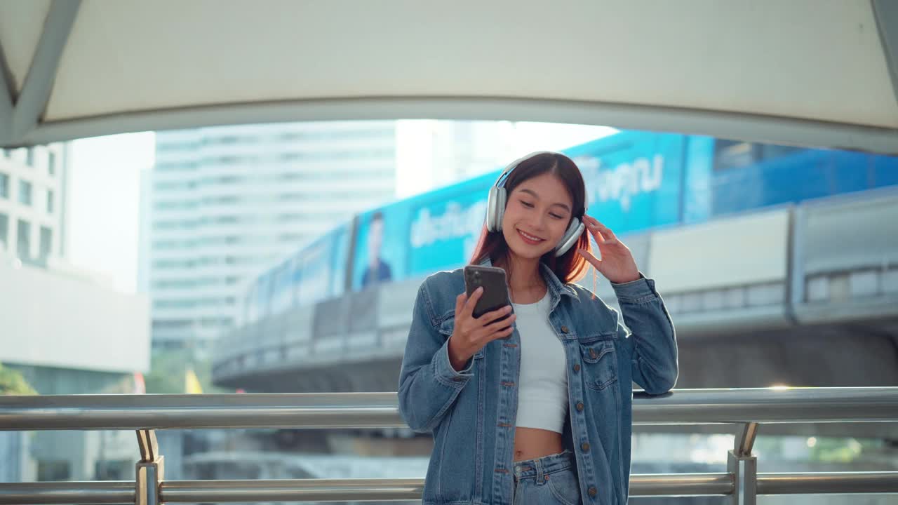 快乐的年轻亚洲女子戴着耳机在的街道上欣赏音乐，微笑的女性独自听音乐跳舞。