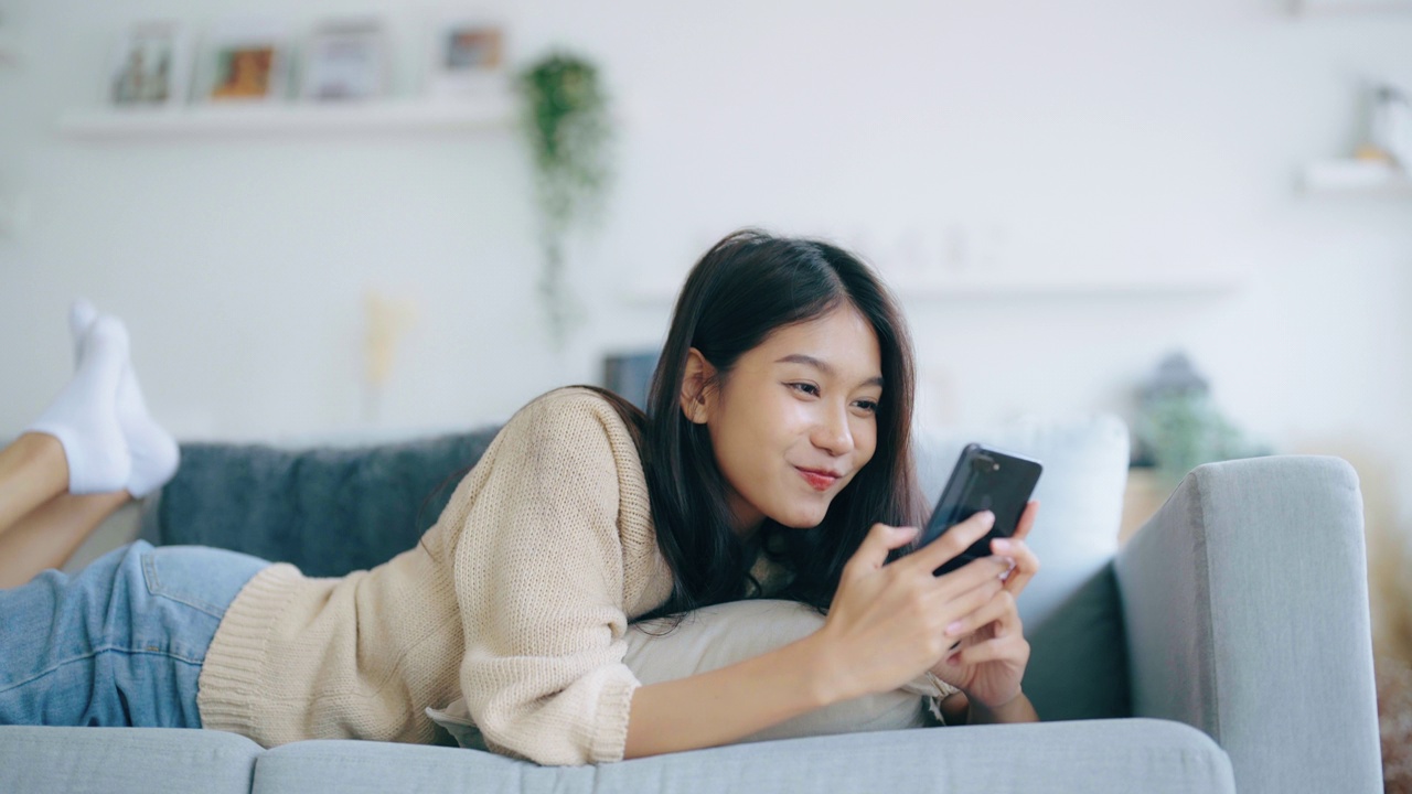 快乐的年轻亚洲女性在家里放松。女人笑着躺在沙发上，手里拿着手机。女孩用视频打电话给朋友