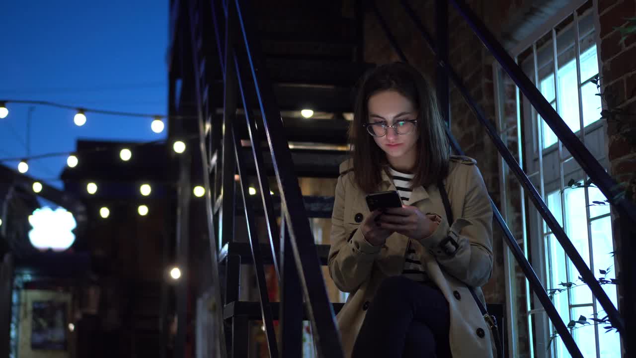 一名年轻女子坐在防火梯上用智能手机发短信。深夜，一个戴眼镜的女孩站在狭窄的街道上，手里拿着电话。4 k