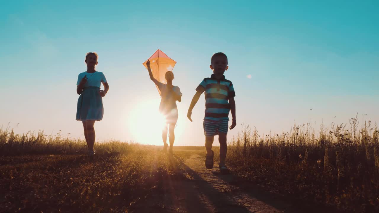 快乐的年轻妈妈带着孩子们一起在公园里放风筝。男孩手里拿着风筝在跑。夕阳下公园里快乐的人们奔跑的剪影。幸福父母童年梦想概念。
