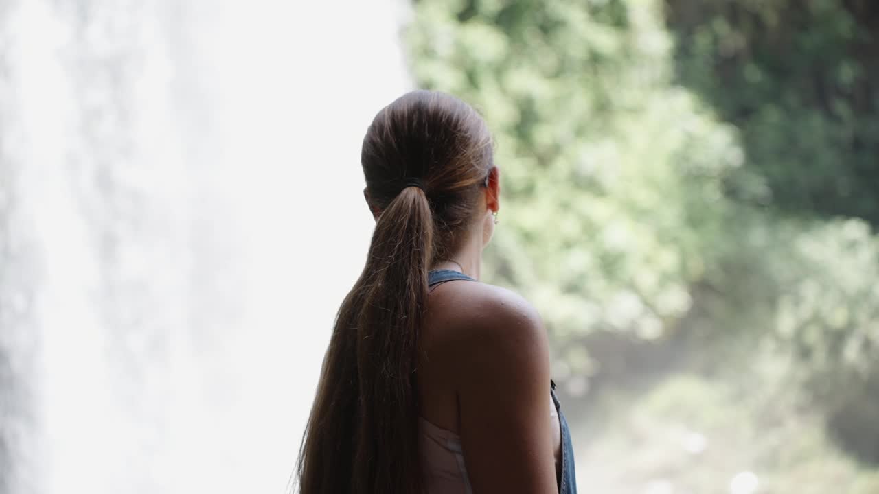 一位身穿牛仔连衣裙的年轻女子正在欣赏森林中山间河流中的瀑布。