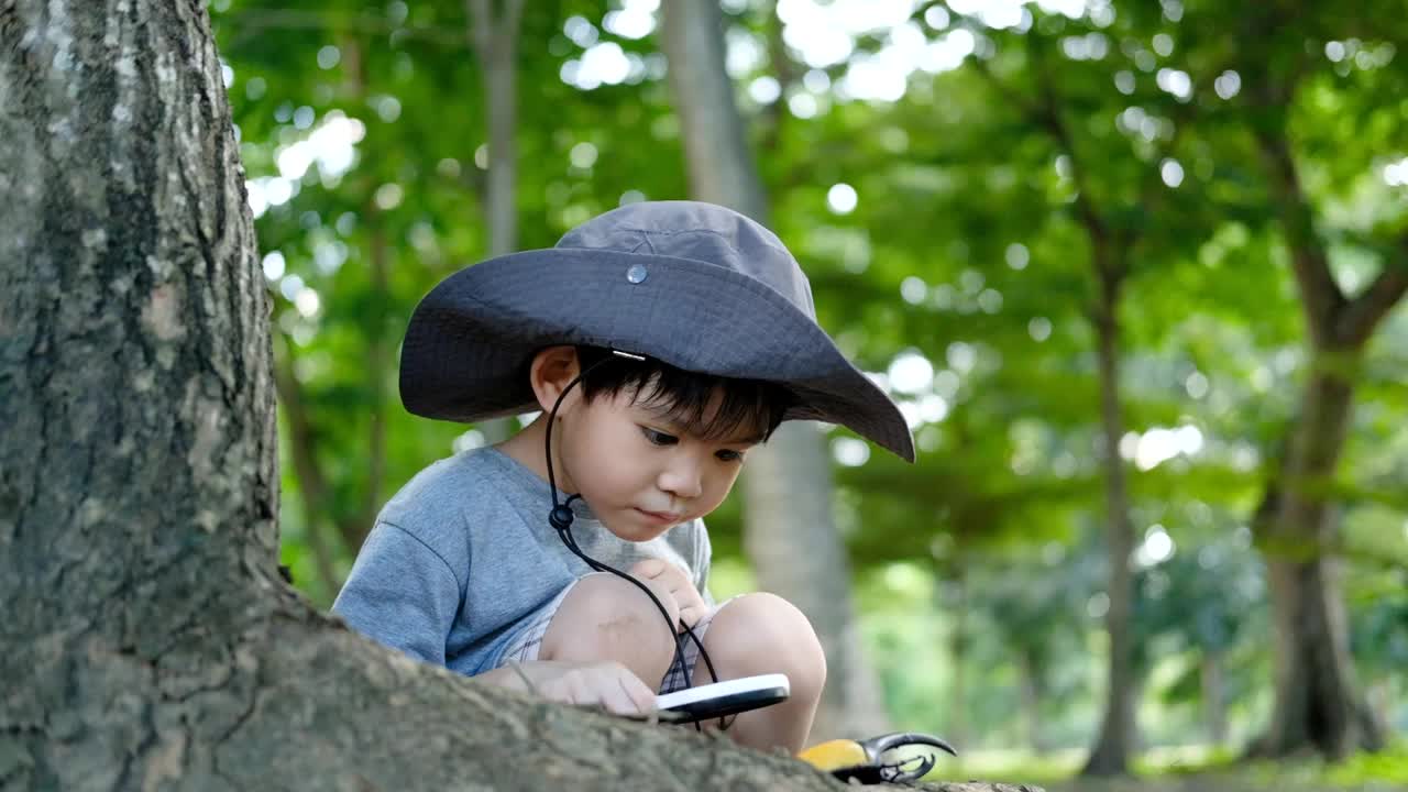 一个亚洲男孩戴着帽子，穿着森林探险服，玩着手上的甲虫。