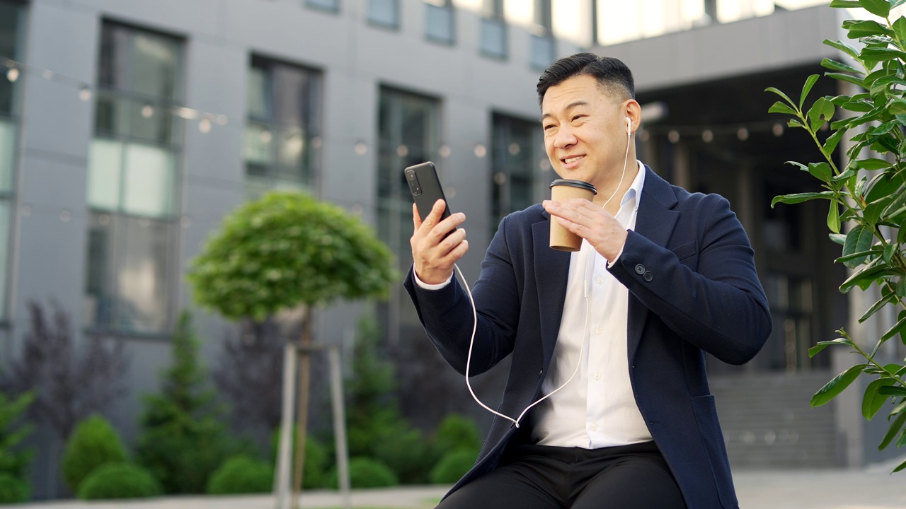 一名身着正装的亚洲商人坐在办公大楼附近街道的长椅上用智能手机进行视频通话。
