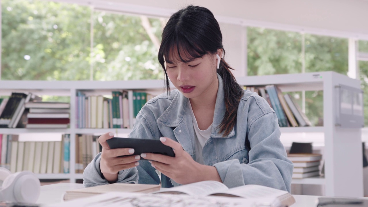 亚洲年轻的女大学生在大学图书馆的课间休息时间用手机玩网络游戏。在学术追求中捕捉悠闲的放松时刻。