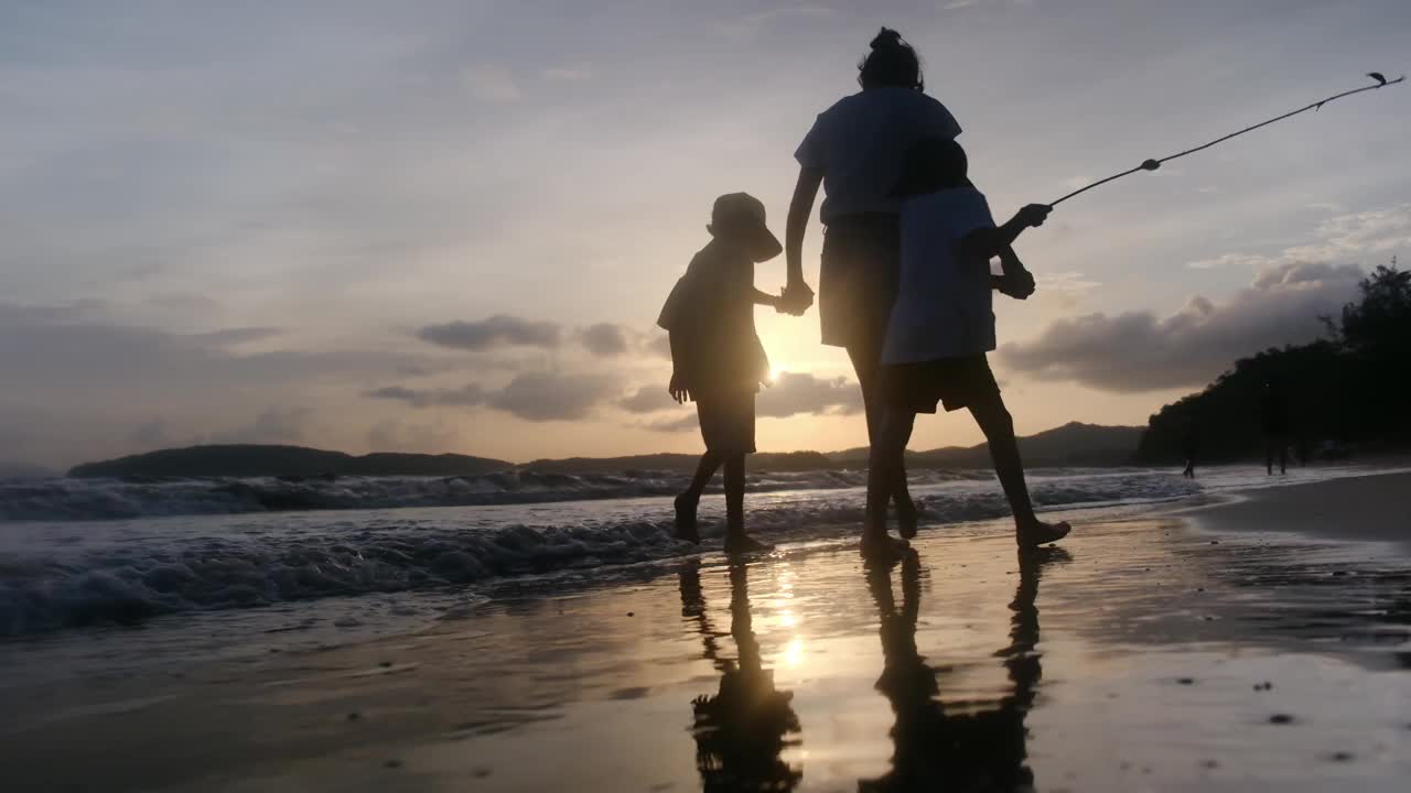 年轻的母亲带着两个儿子在日落的海滩上，夕阳的剪影母子手牵着手看着大海和日落。