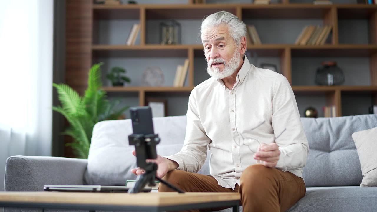 詳情圖-自信的老年白發(fā)男性私人教師坐在沙發(fā)上用智能手機進行視頻通話課程或培訓