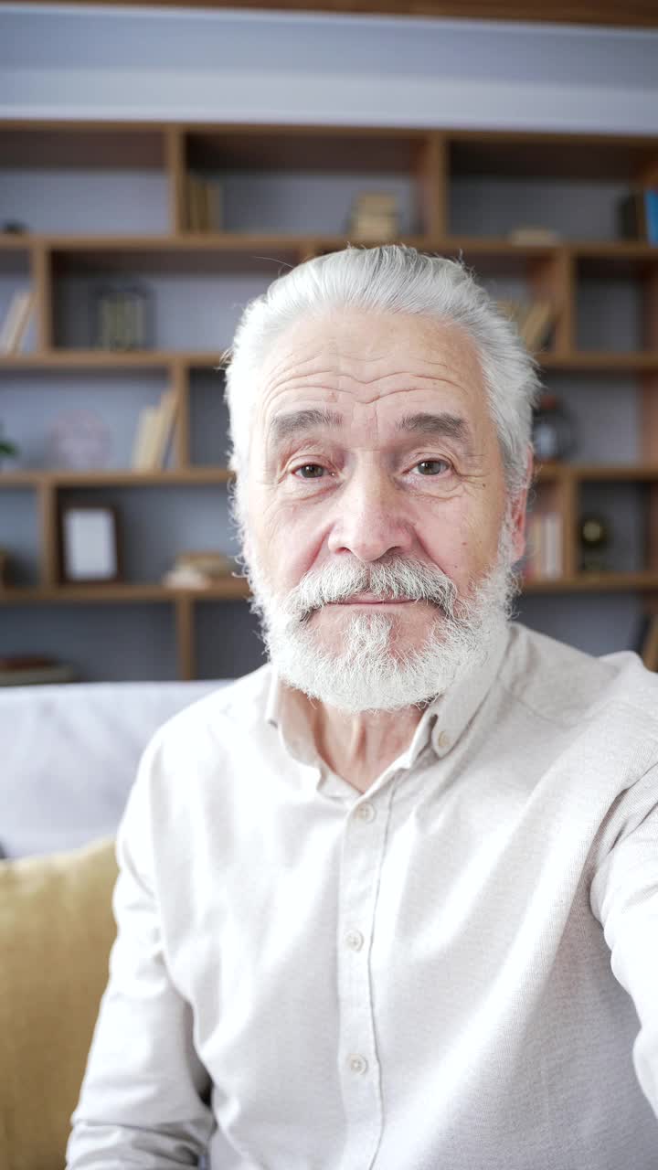 垂直视角:一位白发老人坐在家里客厅的沙发上用智能手机进行视频通话自拍。