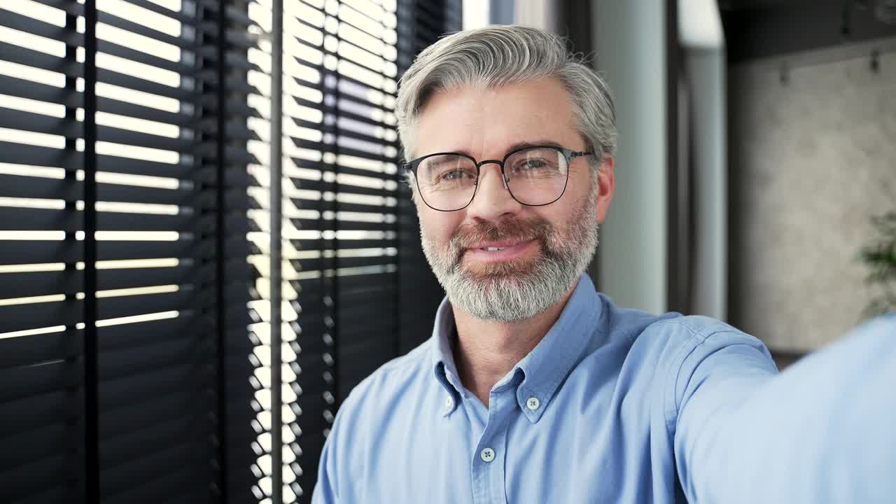 POV一位面带微笑的成熟的白发胡须商人在办公室用智能手机进行视频通话自拍