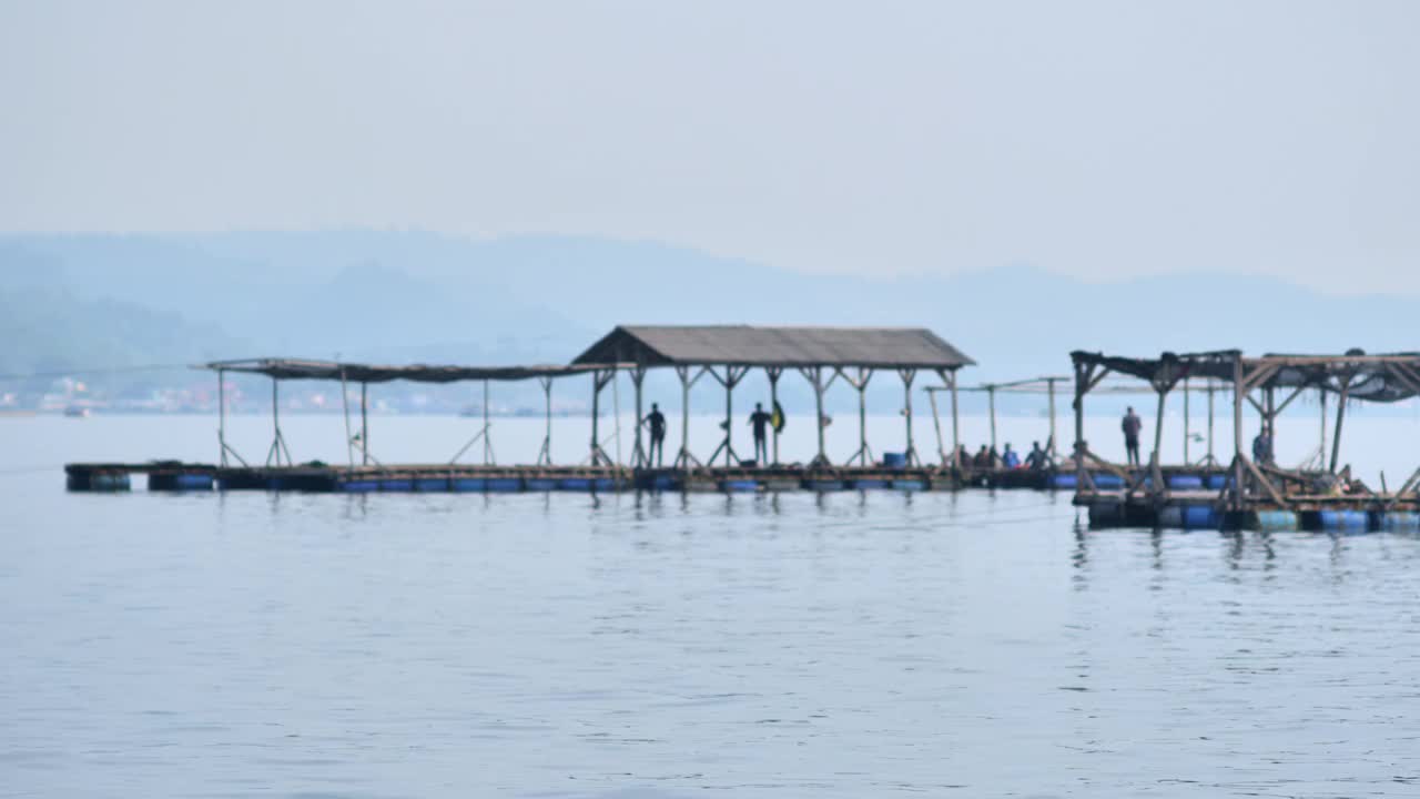 在楠榜平静的海面上，人们在漂浮的笼子上捕鱼，远处是渔村。