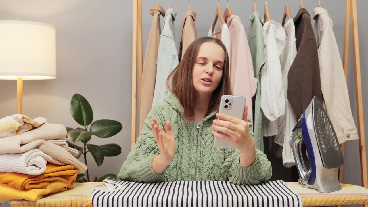 可愛的棕色頭發(fā)的年輕女子穿著針織襯衫在家里熨燙衣櫥里的衣服，在家務休息的時候和朋友視頻通話，閑聊