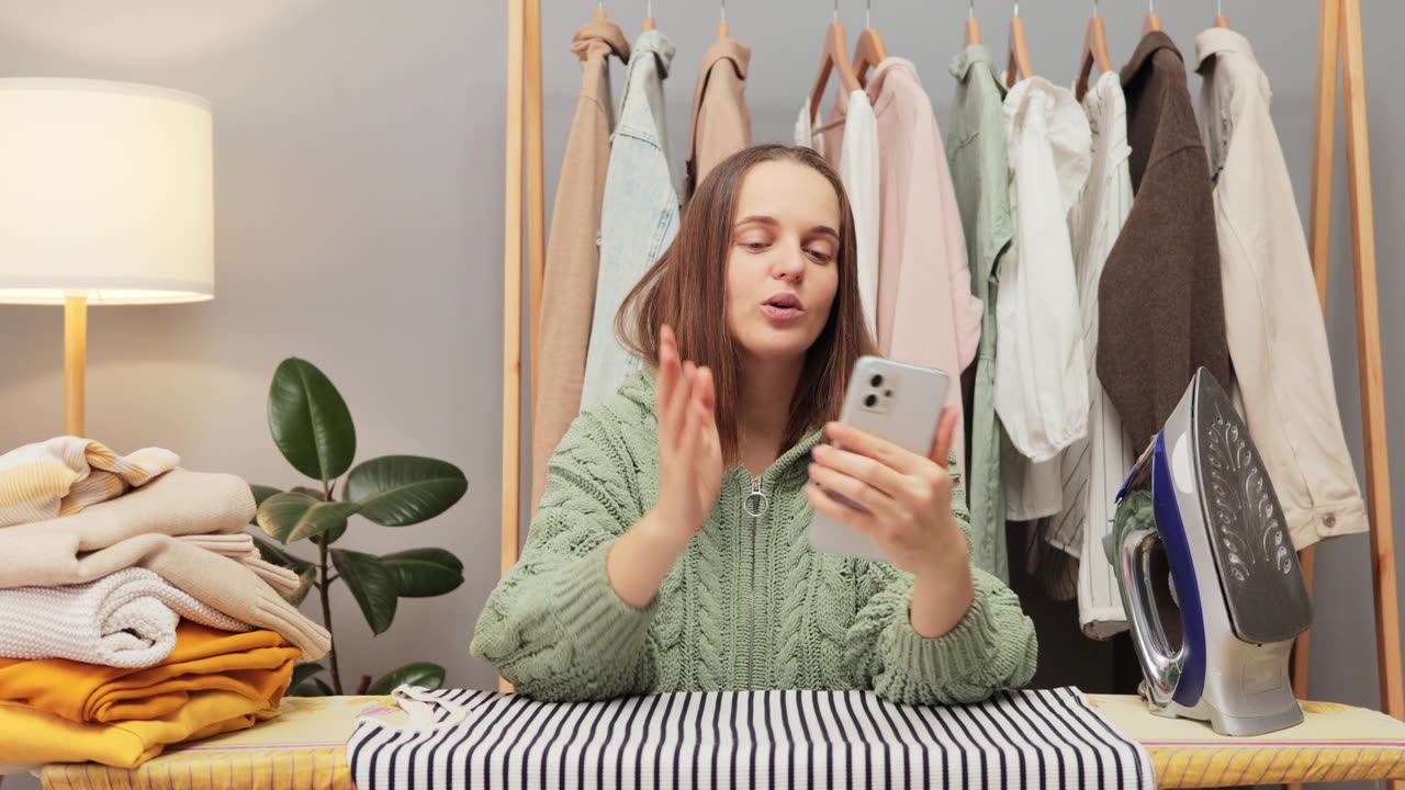 詳情圖-可愛的棕色頭發(fā)的年輕女子穿著針織襯衫在家里熨燙衣櫥里的衣服，在家務(wù)休息的時(shí)候和朋友視頻通話，閑聊