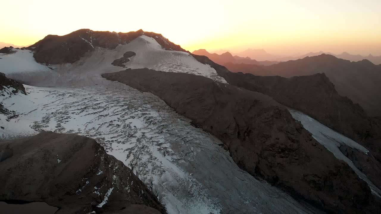 空中俯瞰两名徒步旅行者在瑞士瓦莱州阿罗拉附近的瓦松冰川悬崖上欣赏风景，日落时分，层层叠叠的山脉后面闪烁着五彩缤纷的光芒